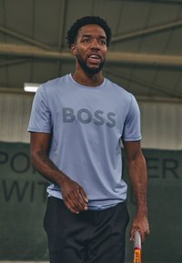 Camiseta atlética de color azul claro con la palabra "BOSS" en franjas oscuras, combinada con pantalones cortos negros y sosteniendo una raqueta de tenis.