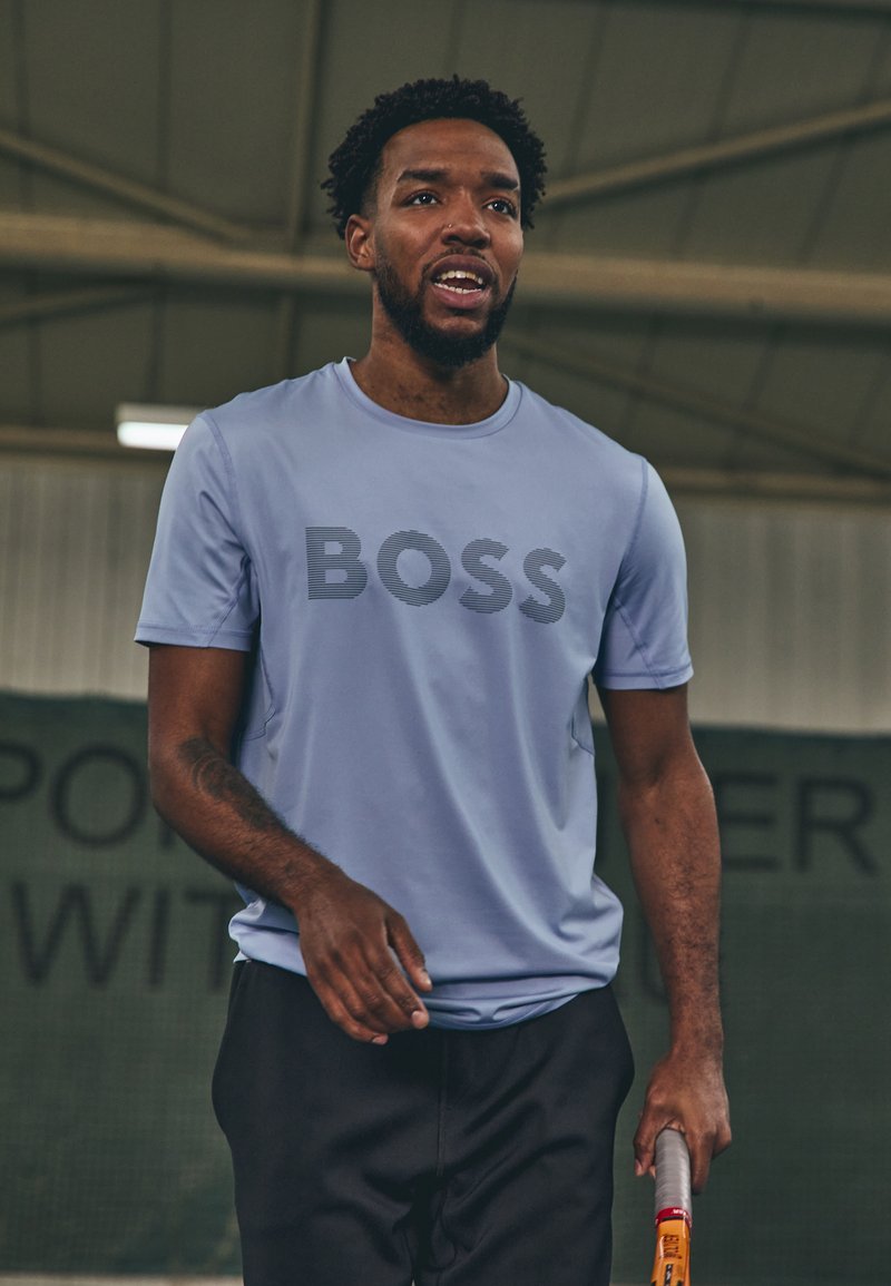 Camiseta atlética de color azul claro con la palabra "BOSS" en franjas oscuras, combinada con pantalones cortos negros y sosteniendo una raqueta de tenis.