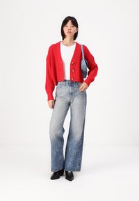 Rode gebreide cardigan met drie knopen, een wit shirt eronder, gecombineerd met lichtblauwe flare jeans met geborduurde sterpatronen en zwarte laarzen.
