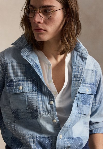 Jeune personne aux cheveux bruns mi-longs portant des lunettes rondes, une chemise blanche et une veste à boutons patchwork bleue.