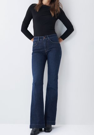 Kvinde iført mørkeblå højtaljede flare-jeans, sort langærmet top og sorte ankelstøvler, stående mod en ensartet lys baggrund.