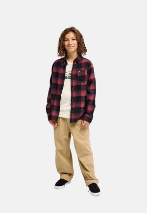 Enfant aux cheveux bouclés portant une chemise à carreaux rouge et noire, pantalon beige, chaussures noires et blanches, debout devant un fond uni.