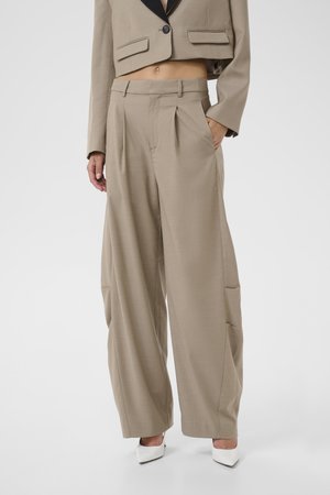 Donna che indossa pantaloni beige a gamba larga con pieghe e tasche laterali abbinati a un blazer beige corto e tacchi bianchi appuntiti.