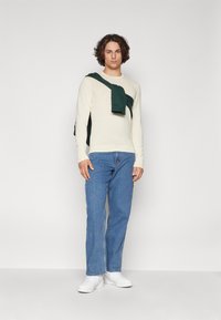 Cream sweater worn over the shoulders, blue denim jeans, and white sneakers. The outfit features a relaxed fit and smooth textures.