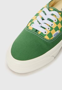 Green canvas sneaker with yellow checkerboard pattern on the toe, white laces, and a textured rubber sole. Features Vans branding.