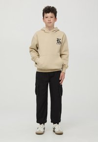 Felpa beige con cappuccio e tasca frontale, con logo nero, abbinata a pantaloni cargo neri con tasche laterali e sneaker bianche.