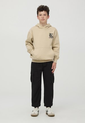 Felpa beige con cappuccio e tasca frontale, con logo nero, abbinata a pantaloni cargo neri con tasche laterali e sneaker bianche.