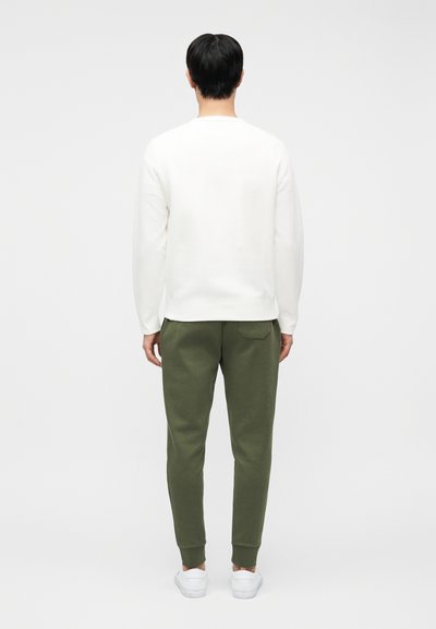 Άνδρας που στέκεται με την πλάτη προς την κάμερα φορώντας άσπρη μπλούζα με μακριά μανίκια, χακί παντελόνι jogger και άσπρα αθλητικά παπούτσια σε απλό φόντο.