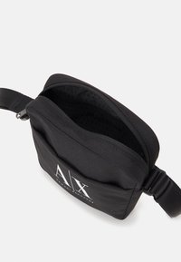 Czarna torba na ramię z tkaniny z białym logo "A|X Armani Exchange", częściowo otwarty suwak i regulowany pasek na białym tle.