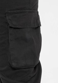 Poche de pantalon cargo noir avec fermeture à rabat, montrant la texture du tissu et le détail de couture sur le côté de la jambe.