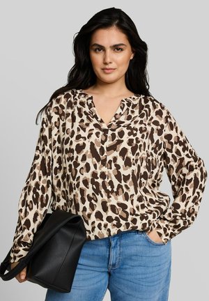 Vrouw met lang donker haar, die een beige en bruine blouse met luipaardprint en blauwe spijkerbroek draagt, houdt een zwarte leren tas in één hand.