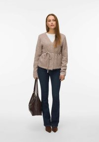 Svijetlosmeđi pleteni kardigan s vezom u struku, dugim rukavima i V-izrezom, nošen preko bijele majice, upotpunjen tamnim trapericama i smeđom pletenom torbom.