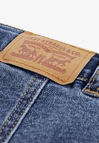 Smeđa kožna oznaka s brendom s utisnutim dizajnom, pričvršćena na plavu traper tkaninu, s žutim šavovima i teksturnim detaljima.