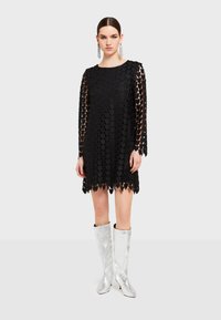 Musta mekko, jossa on teksturoitu muotoilu, jossa on päällekkäisiä sydämenmuotoisia palasia, pitkät hihat ja aaltoileva helma. Yhdistetty kiiltäviin hopeisiin saappaisiin.