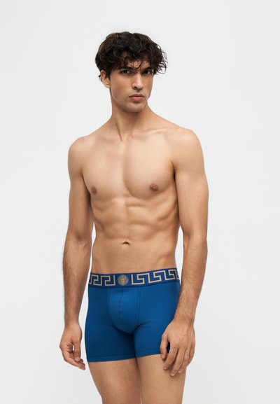 Jeune homme aux cheveux bouclés portant un boxer bleu avec une ceinture dorée à motifs, debout devant un fond clair uni.