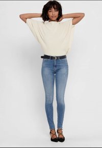 Haut crème oversized à manches courtes, associé à un jeans skinny bleu clair. Une ceinture noire et des talons aiguilles noirs complètent le look.