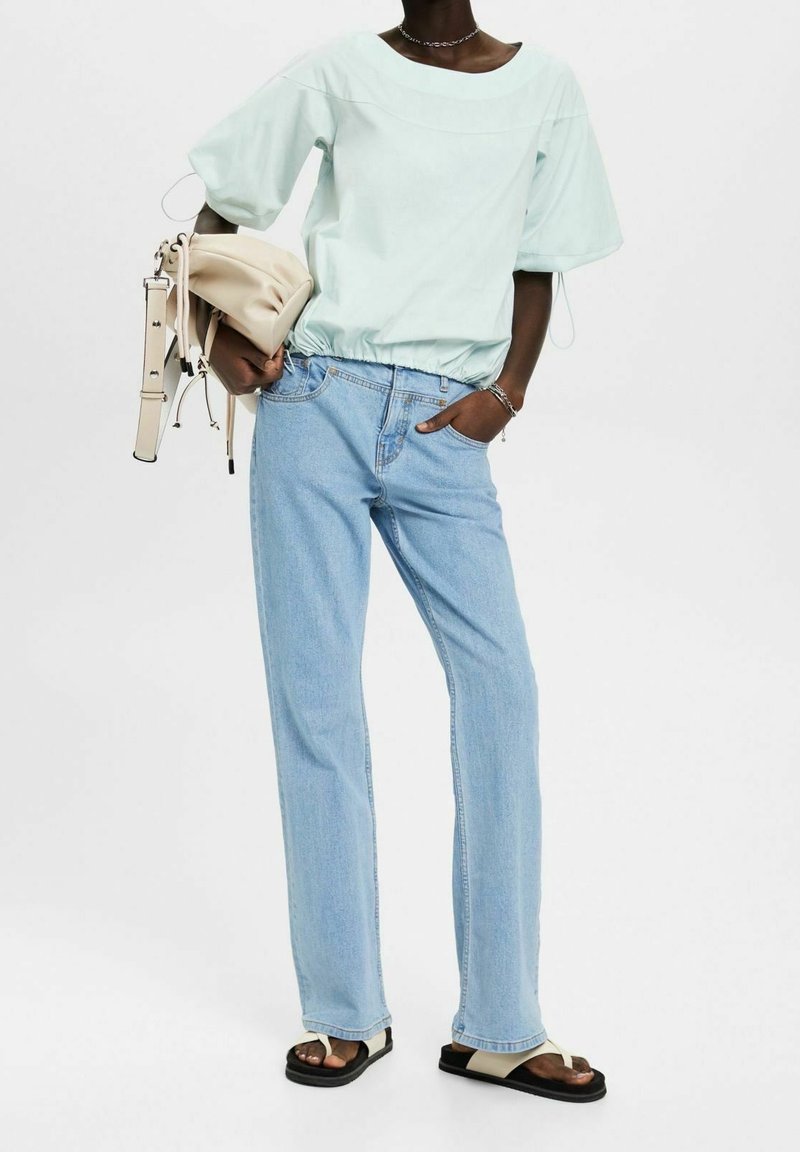Personne portant un top à manches bouffantes bleu clair, un jean droit bleu clair, des sandales beige, tenant un sac à main beige avec une bandoulière sur fond blanc.