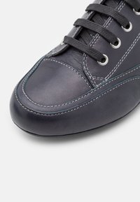 Gros plan d'une chaussure en cuir gris foncé avec des lacets noirs et des coutures blanches visibles sur un fond clair uni.