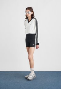 Young person wearing white long-sleeve crop top, black high-waisted shorts with stripes, white socks, gray sneakers, and sunglasses standing on blue floor.