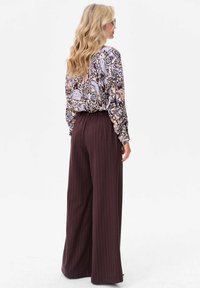Bloes met bloemmotief in tinten blauw, perzik en crème; hoog nazittende bruine broek met streepjes en wijde pijpen; model gezien vanuit de rug.