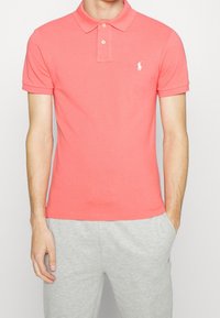 Korallfärgad polo med korta ärmar, tillverkad av bomull, med knäppning med tre knappar och liten broderad logotyp på bröstet. Matchas med gråa sweatpants.