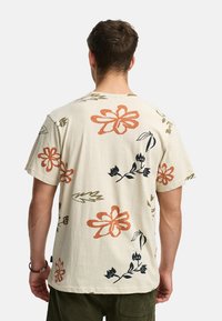 Camiseta de manga corta en un tono beige claro con diseños florales en naranja, negro y verde. Textura suave con un corte holgado.