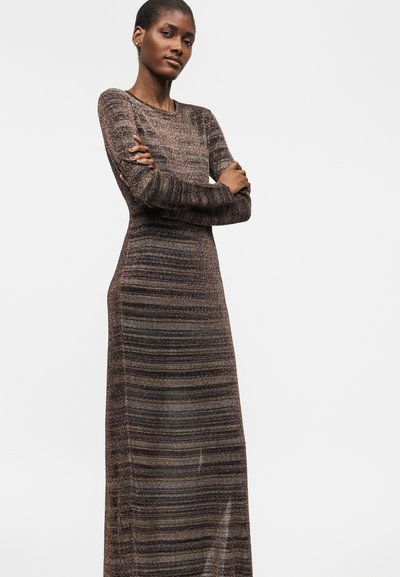 Γυναίκα με σταυρωμένα χέρια που φοράει ένα πλεκτό φόρεμα μακρυμάνικο, μακρύ έως το πάτωμα, με οριζόντιες ρίγες σε αποχρώσεις του καφέ και του γκρι.