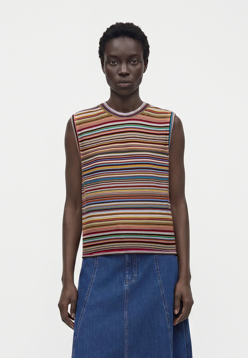 Femme aux cheveux courts et bouclés portant un haut sans manches rayé multicolore et une jupe en jean bleue, sur fond blanc uni.