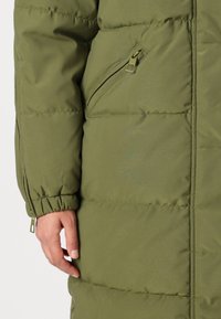 Olive green puffer coat with a textured fabric, featuring a zippered pocket, elastic cuff detailing, and a quilted pattern.