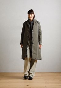 Khaki lange jas met knopen, gedragen over een donkere top en lichte wijde broek. Soepele textuur, eenvoudig ontwerp en rechte silhouet.