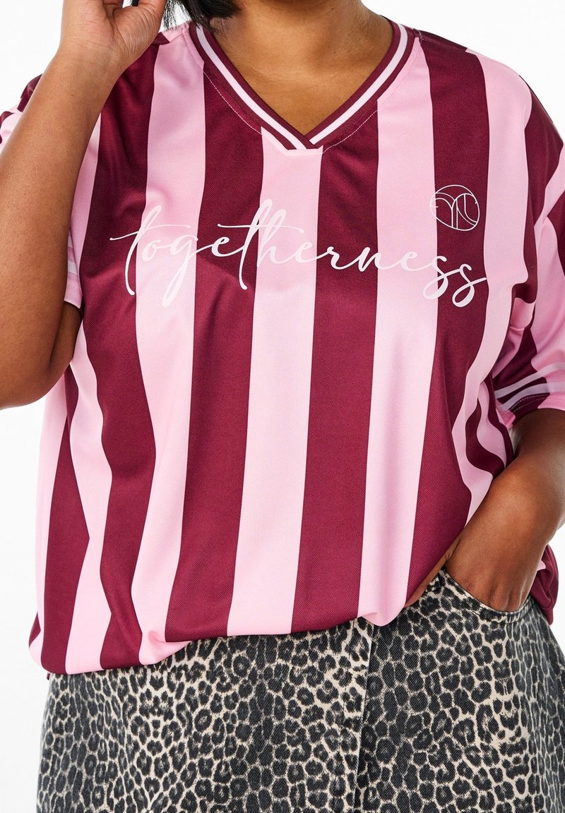 Personne portant une chemise rayée bordeaux et rose avec le texte "togetherness" et un pantalon à motif léopard, main dans la poche reposant sur la hanche.