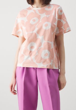 Femme portant une chemise à manches courtes ample avec de grandes impressions de fleurs de pêche et un pantalon plissé taille haute de couleur violet vif.