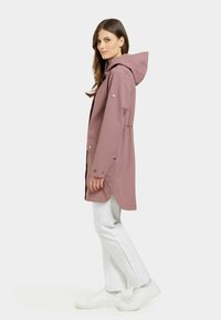 Rosa vattentät jacka med huva, dragsko i midjan, tryckknappar och sidofickor. Bärs över vita byxor och vita sneakers.