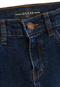 Jeans in denim blu scuro con etichetta Guess, bottone di rame e cuciture arancioni a contrasto lungo le cuciture e le tasche.