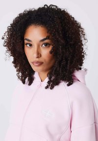 Haut à zip rose avec une texture douce, arborant un logo et des accents blancs. Le design comprend une capuche et des poches kangourou.