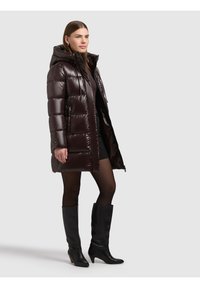 Brūna pūkaina virsjaka ar kapuci, spīdīga auduma, šūta dizaina un priekšā ar rāvējslēdzēju. Valkāta virs melnas kleitas un kopā ar ceļgalus sasniedzošām melnām zābakiem.