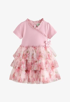Junges Mädchen trägt ein rosa Kleid mit Rüschen und floralen Tüllschichten und weiße Sneakers mit großen rosa Schleifen, steht auf grauem Boden.