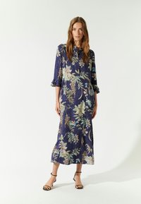 Langes, marineblaues Blumenkleid mit Dreiviertelärmeln und Rüschenbündchen, das hellfarbige botanische Muster aufweist. Getragen mit schwarzen Sandalen mit Riemen.