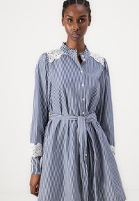 Dryžuota suknelė tamsiai mėlynos ir baltos spalvos, pagaminta iš medvilnės. Suknelė turi nėriniuotas detales pečių ir rankovių srityse, sagų priekį, ir juosmens diržą. A linijos siluetas.