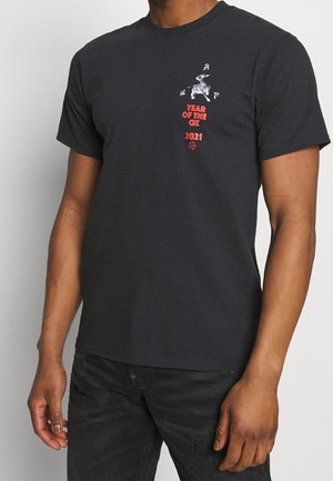 Personne portant un t-shirt noir avec un petit bœuf argenté et un texte rouge « Année du Bœuf 2021 » sur le haut gauche de la poitrine.