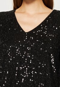 Femme portant un haut noir à col en V recouvert de petits sequins noirs brillants sur un fond neutre.