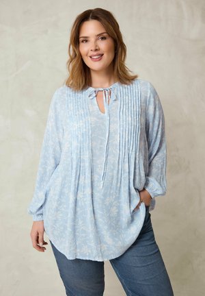 Nainen pukeutunut vaaleansiniseen kukalliseen pitkähihaiseen puseroon, jossa on laskosyksityiskohtia, ja farkkuihin. Hän seisoo, toinen käsi taskussa, hymyillen.