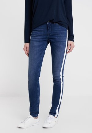 Persoon draagt donkere blauwe skinny jeans met witte zijstrepen, een marineblauwe lange mouwen top en witte sneakers, staand tegen een effen achtergrond.