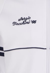 Witte sportjasje met marineblauwe accenten. Bevat geborduurde tekst "Sergio Tacchini '86" en twee marineblauwe strepen op de mouwen. Gladde textuur.