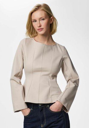 Plavuša odjevena u tijesnu bež majicu dugih rukava i tamnoplave traperice, pozira s rukama u džepovima na jednobojnoj pozadini.