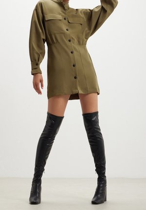 Person wearing olive green button-up mini dress and black knee-high heeled boots, standing with one arm raised.