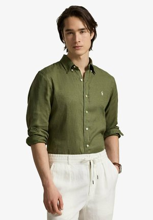 Jeune homme portant une chemise boutonnée vert olive avec manches retroussées et un pantalon blanc à cordon, debout avec une main dans la poche.