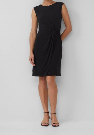 Kobieta w czarnej, bezrękawnej sukience do kolan z wiązaniem po boku oraz w czarnych, wysokich sandałach na paski, stoi na tle białej ściany.