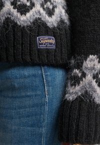 Close-up van een zwart-wit gebreide trui met patroon en een Superdry-label, gecombineerd met blauwe spijkerbroek.