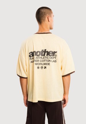 Lyhyttukkainen mies, joka pukeutunut löysään beigeen T-paitaan, jossa on mustaa tekstiä ja kuvakkeita selässä, yhdistettynä tummiin housuihin.